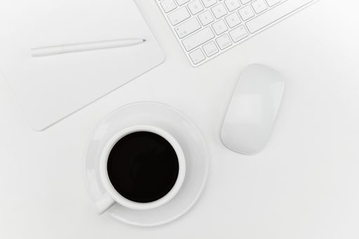 A top view of a cup of coffee, notebook, and a keyboard and mouse on a white desk