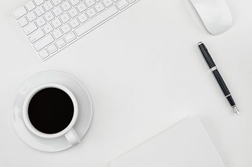A top view of a cup of coffee, pen, and keyboard and mouse on a white desk