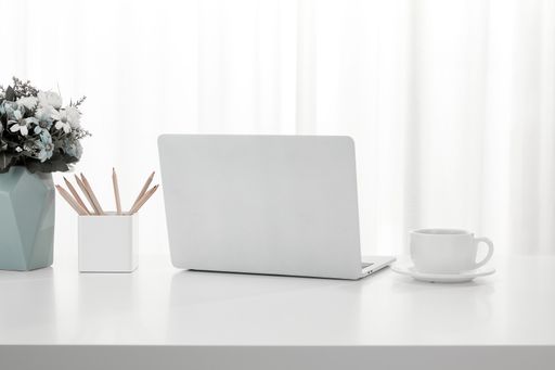 A closeup of a cup of coffee and laptop on a white desk, next to a pencil holder and vase full of flowers
