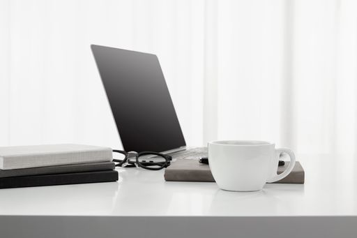 A closeup of a cup of coffee, laptop, notebooks, and a pair of glasses on a white desk