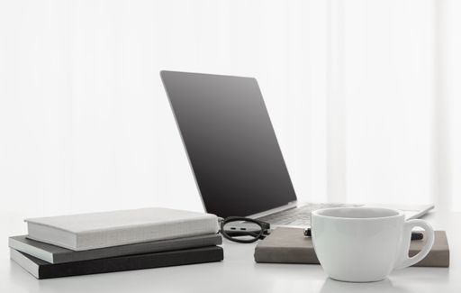 A closeup of a cup of coffee, laptop, notebooks, and a pair of glasses on a white desk