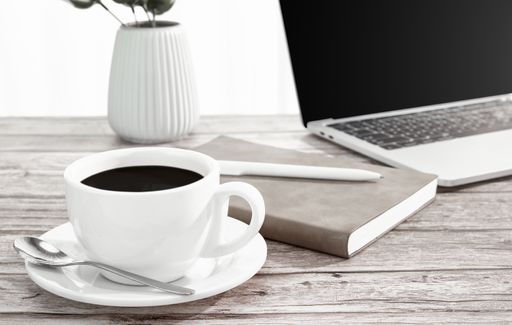 A closeup of a cup of coffee, laptop, notebook, and pen on a wooden surface