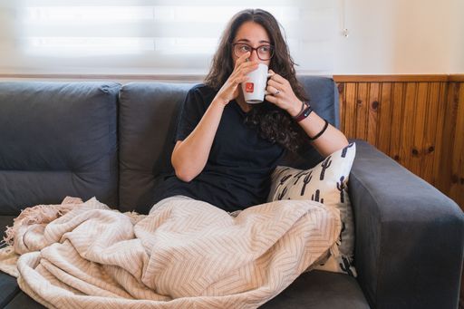 An eye-level shot of a Hispanic woman sitting on a sofa with a soft blanket and drinking a hot tea
