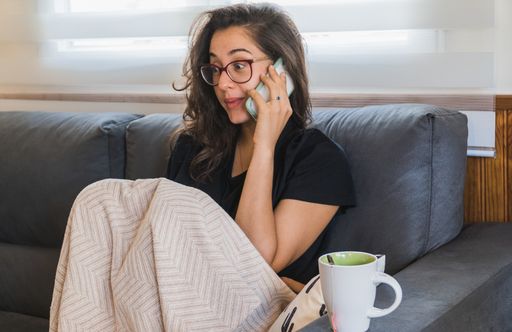 An eye-level shot of a cute attractive Hispanic woman sitting on a sofa with a blanket and cup of tea while talking to the phone