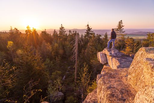 A male hiker standing on the edge of the rocky mountain and enjoying the magical sunset view in the forest
