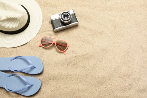 A top view of a camera, sunglasses, slippers and a hat placed on the sand on the beach
