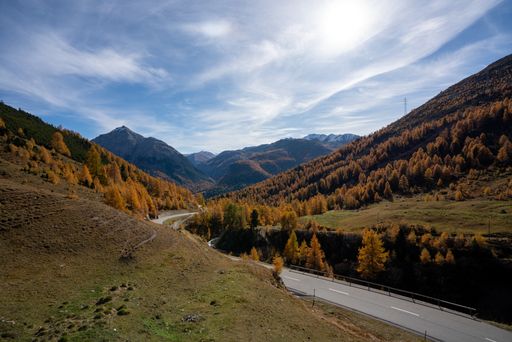 A road surrounded by hills covered in trees under a blue cloudy sky in autumn