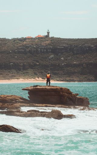A fisherman on rocky formation in Palm Beach, Sydney, NSW, Australia with a lighthouse and headland