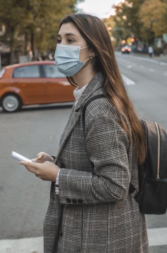A young caucasian brunette female in a face mask and a checkered jacket using her mobile phone while walking in the street