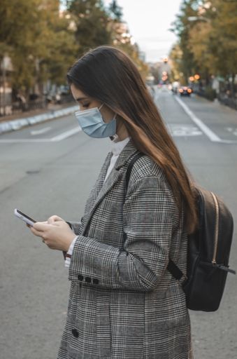 A young caucasian brunette female in a face mask and a checkered jacket using her mobile phone while walking in the street