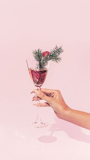 A female hand holds a champagne glass with a Christmas tree branch and a red ball as decoration on pink background