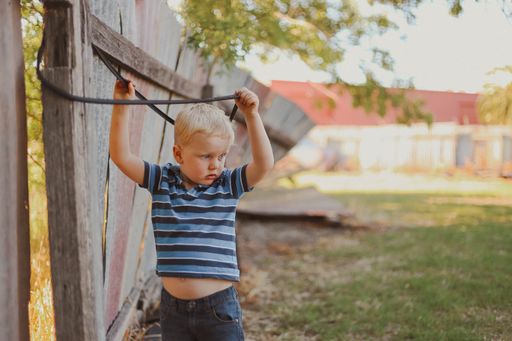 A blonde little boy playing with a rope hanging from an old wooden fence