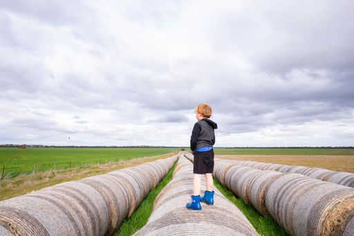 A blonde child standing on a long row of round hay bales on overcast day - childhood on the farm