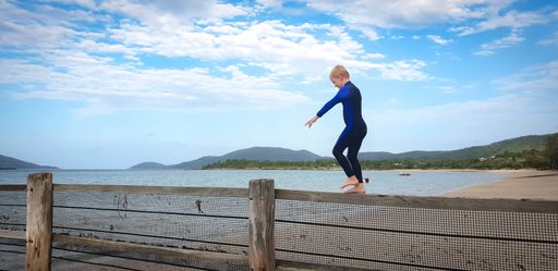 A blonde child wearing a swimming suit and walking on a wooden fence near the beach