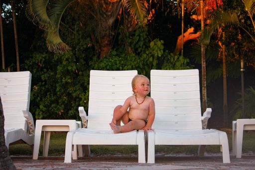 A cute blonde child sitting on a plastic chair near the pool on the background of the trees