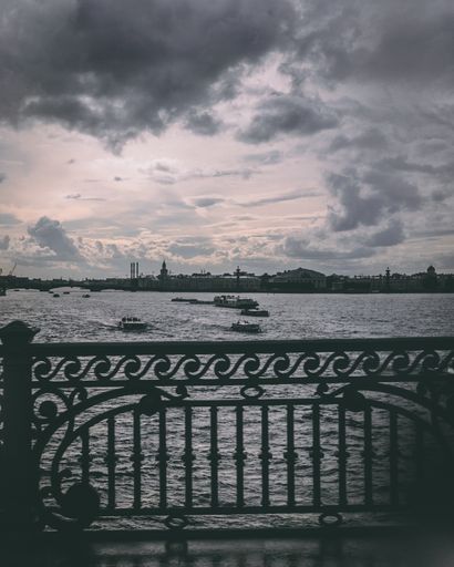 A grayscale vertical shot of boats sailing on a lake under the  cloudy sky  with buildings in the distance
