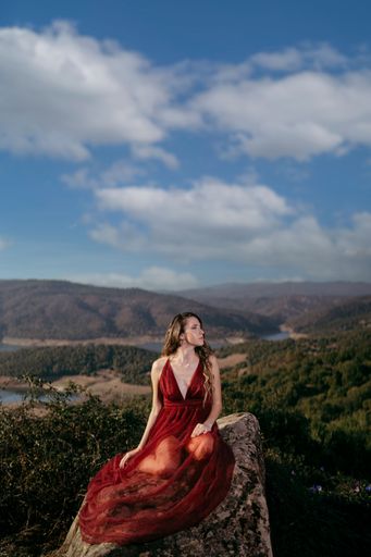 A vertical shot of an attractive female in a long red dress sitting on a rock