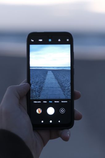 A vertical shot of a hand holding a phone to take a picture of a trail along the sea