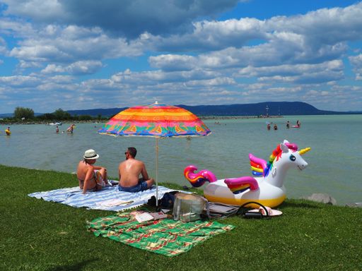 Holiday couple sitting relaxed under a colorful sunshade parasol next to a colorful unicorn on the grassy beach strand of the lake Balaton in Hungary