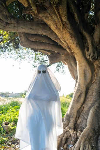 A vertical shot of a person dressed as a ghost standing next to the tree on the day of dead in Mexico