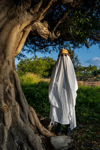 A vertical shot of a person covered by a white sheet, with sunglasses and hat, in appearance of a ghost, in a garden during the day in Mexico