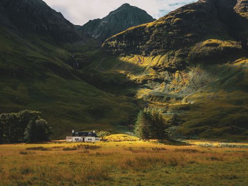 A white house in a field with a black roof surrounded by high and beautiful mountains in Glencoe, Scotland