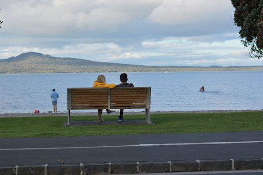 View of man and woman sitting on park bench at Tamaki Drive Mission Bay beach with Rangitoto volcano island