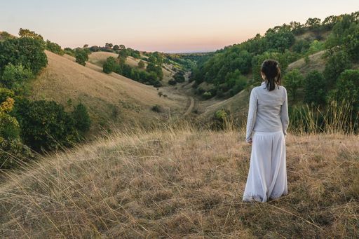 A female in a white dress admiring the horizon while standing in on the dry grass in the mountains