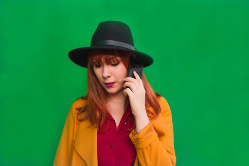 A closeup shot of a redhead lady talking on the phone standing in front of a green wall