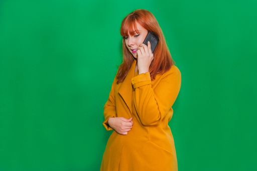 A closeup shot of a redhead lady talking on the phone standing in front of a green wall