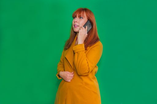 A closeup shot of a redhead lady talking on the phone standing in front of a green wall