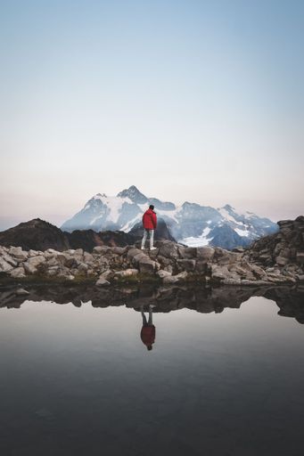 A vertical shot of a man in a red coat enjoying the breathtaking scenery of the Mount Baker at a lake