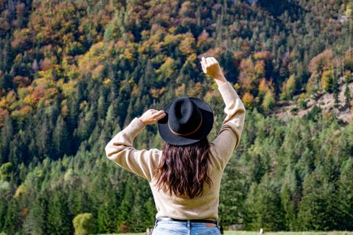 A back view of a young Caucasian woman in a panama hat posing in autumn forest with her arms raised in the air