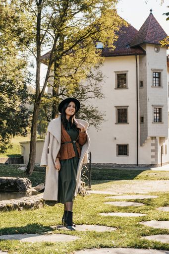 An eye-level vertical shot of an elegant feminine brunette in a stylish coat posing in front of a rural house