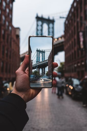 A vertical of a hand holding a phone, taking a photo of Brooklyn Bridge in New York