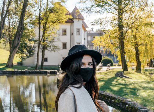 An eye-level closeup portrait of a Caucasian woman in a panama hat and a face mask posing near a park pond