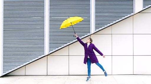 A beautiful blonde female in a purple coat with a yellow umbrella posing in front of a white wall