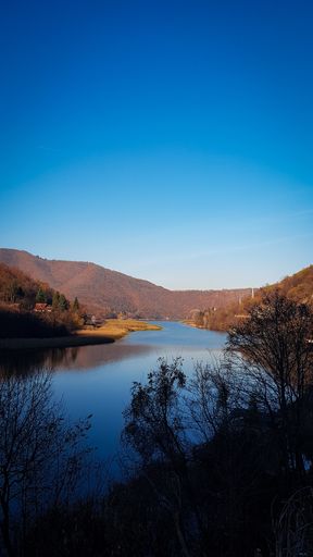A vertical shot of a lake in the middle of forested mountains under a clear blue sky