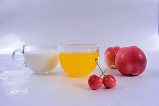 A closeup shot of two glasses with orange juice and milk near peaches and cherries on a white surface