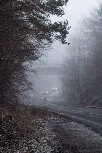 A vertical shot of the road during snowfall with passing cars on a foggy day.