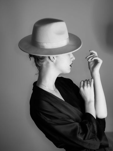 A vertical grayscale shot of a Caucasian woman in a black dress and a hat posing in front of a wall