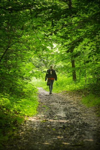 A vertical shot of a woman with gray braided hair walking on a beautiful forest trail surrounded by bright green trees and nature