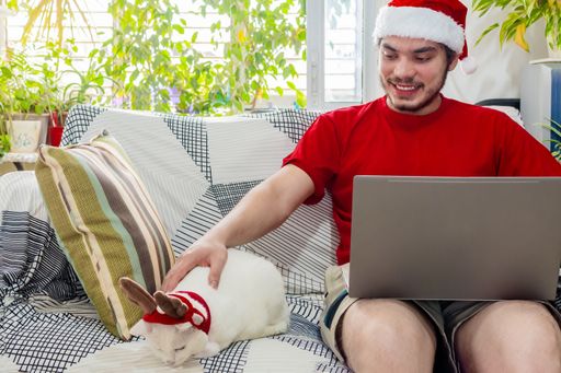 A young Caucasian man in a red shirt and Santa's hat petting his pet cat while working from home during Christmas season