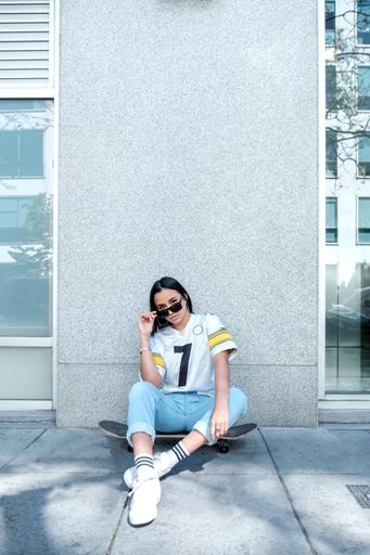 A vertical portrait of a stylish Hispanic woman in sunglasses sitting on a skateboard with an urban building behind