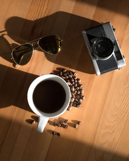 A vertical closeup of a cup of coffee with sunglasses and a retro camera on a wooden table.