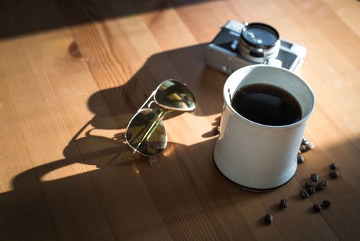 A closeup of a cup of coffee with sunglasses and a retro camera on a wooden table.