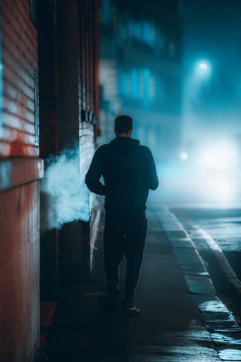 A vertical back view of a man on a sidewalk with smoke and blue lighting in the background