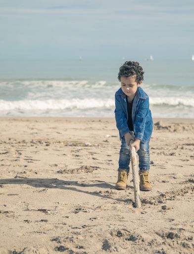 A vertical shot of cheerful little boy in boots playing on the sandy seashore