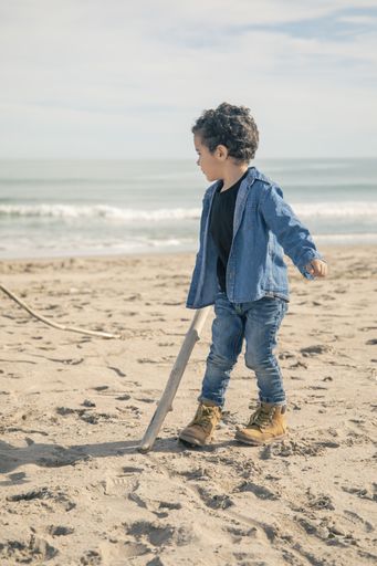 A vertical shot of cheerful little boy in boots playing on the sandy seashore