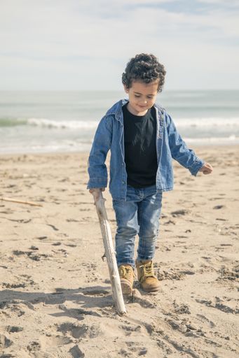 A vertical shot of cheerful little boy in boots playing on the sandy seashore